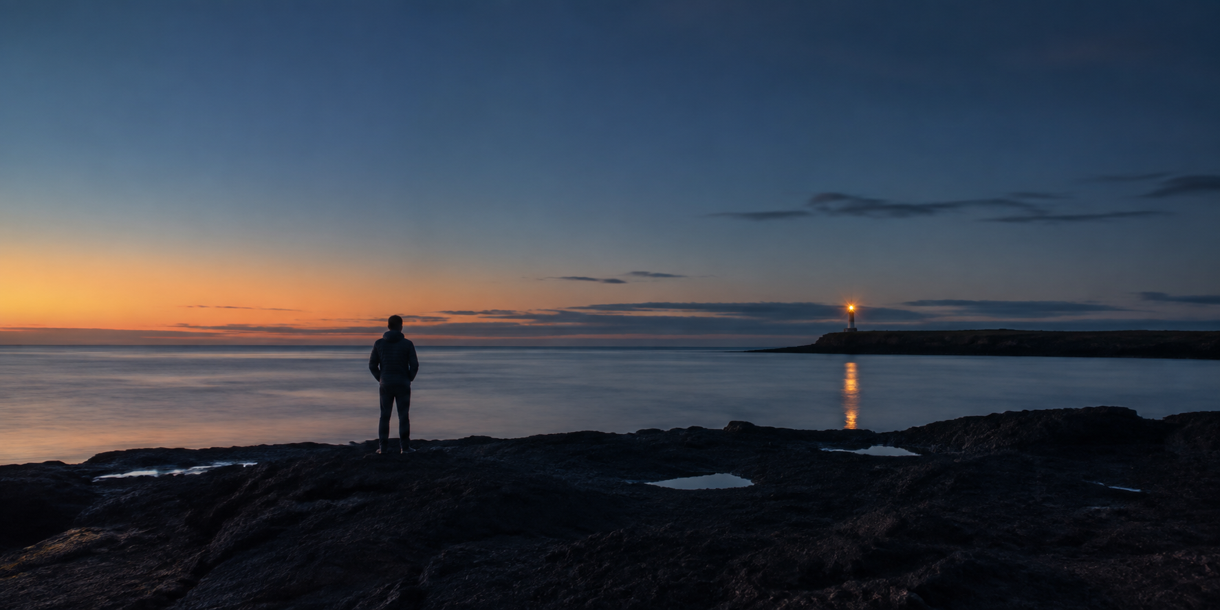 A lone person faces a warm beacon across a calm sea at first light, standing on a dark volcanic shoreline.